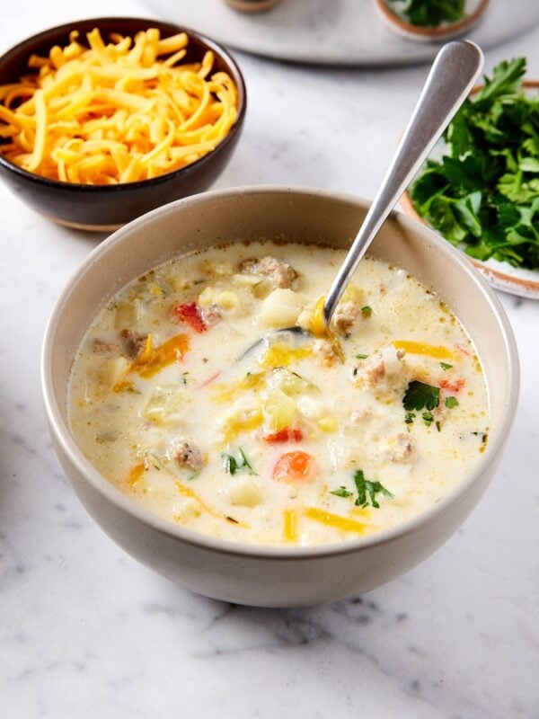 White marble surface with bowl of creamy corn chowder with spoon inside with melted cheese on top and more cheese in background.