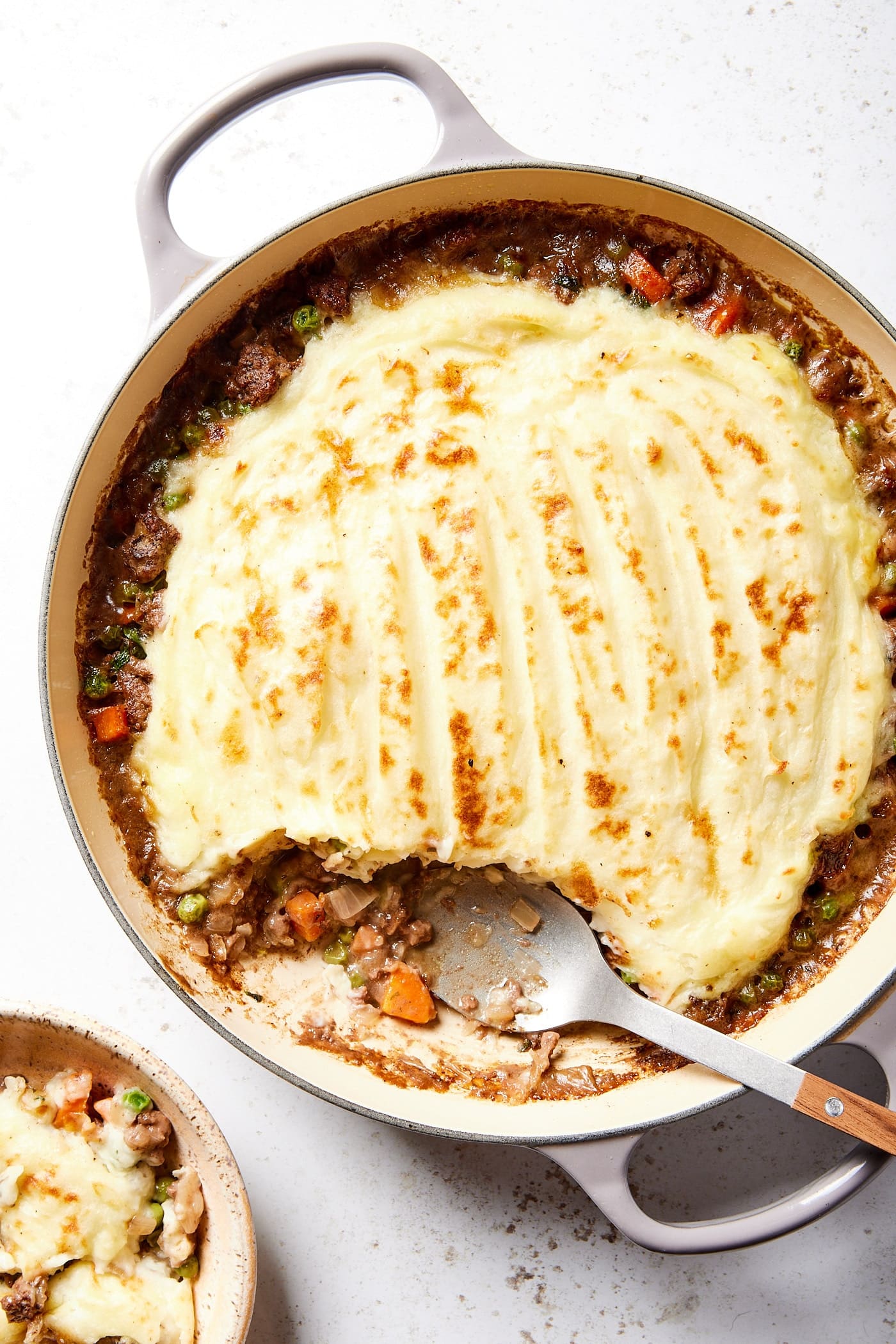 Serving spoon resting against the side of a large pot filled with shepherd's pie with mashed potatoes on top with some in a separate bowl as a serving.