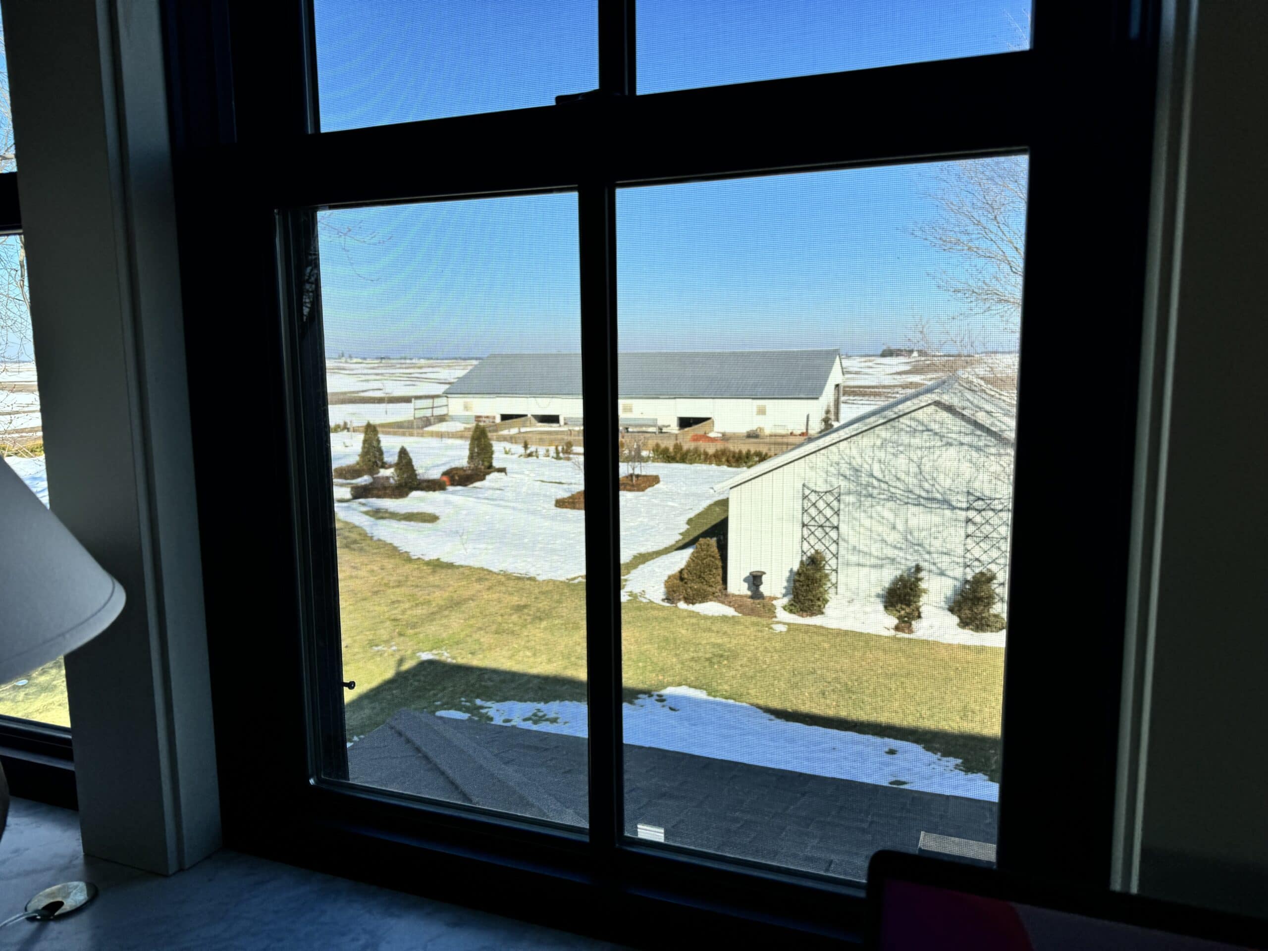 Window looking out from and office onto a backyard with snow on the ground and brown grass.