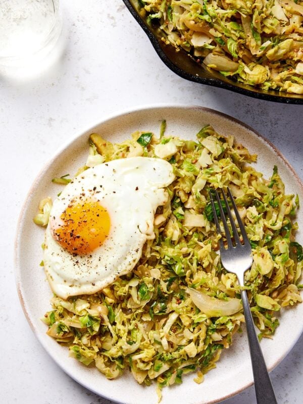 Plate filled with Brussels sprouts has with fried egg to the side with rest of container filled with with the hash as well.