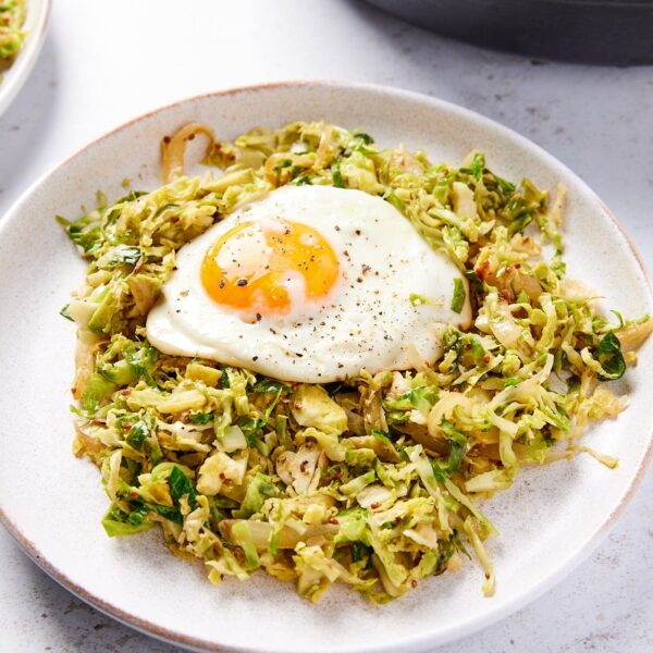 White plate filled with shaved Brussels sprouts has with fried egg on top with skillet in background.