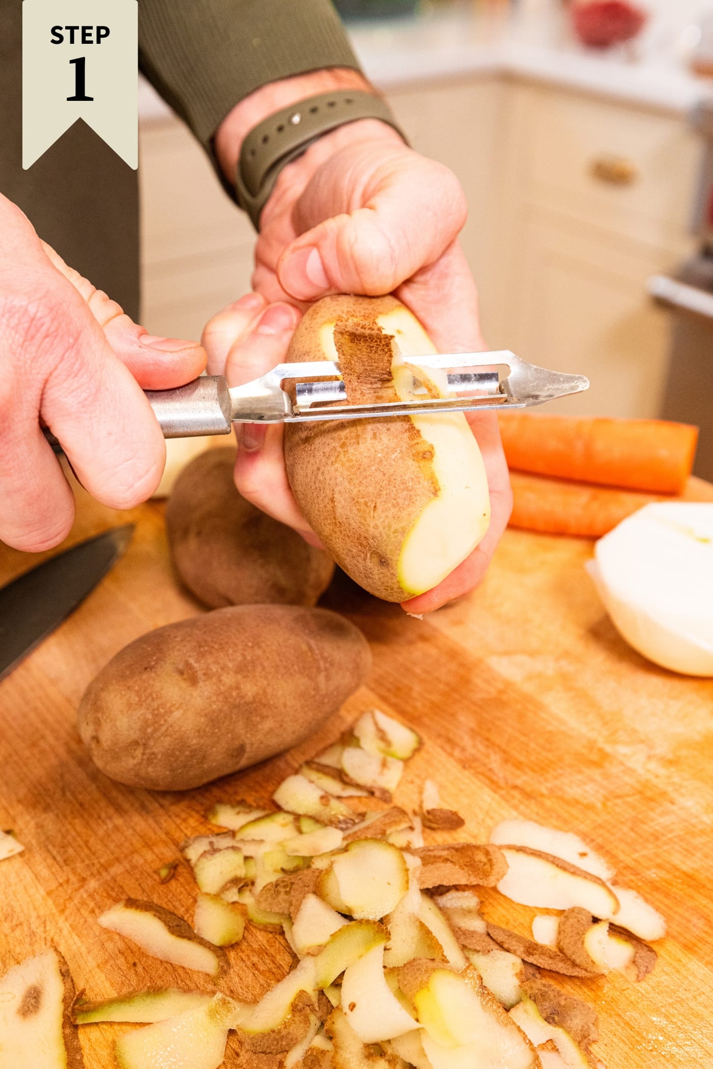 Hand using peeler to peel skin off of potatoes on a wooden cutting board.