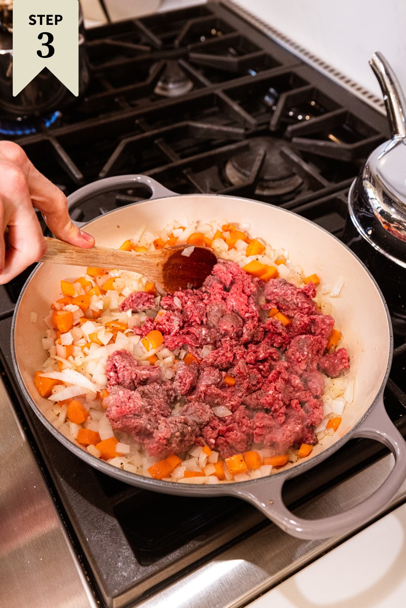Raw ground beef after being added to a large pot on the stovetop with diced carrots and onions already cooking.