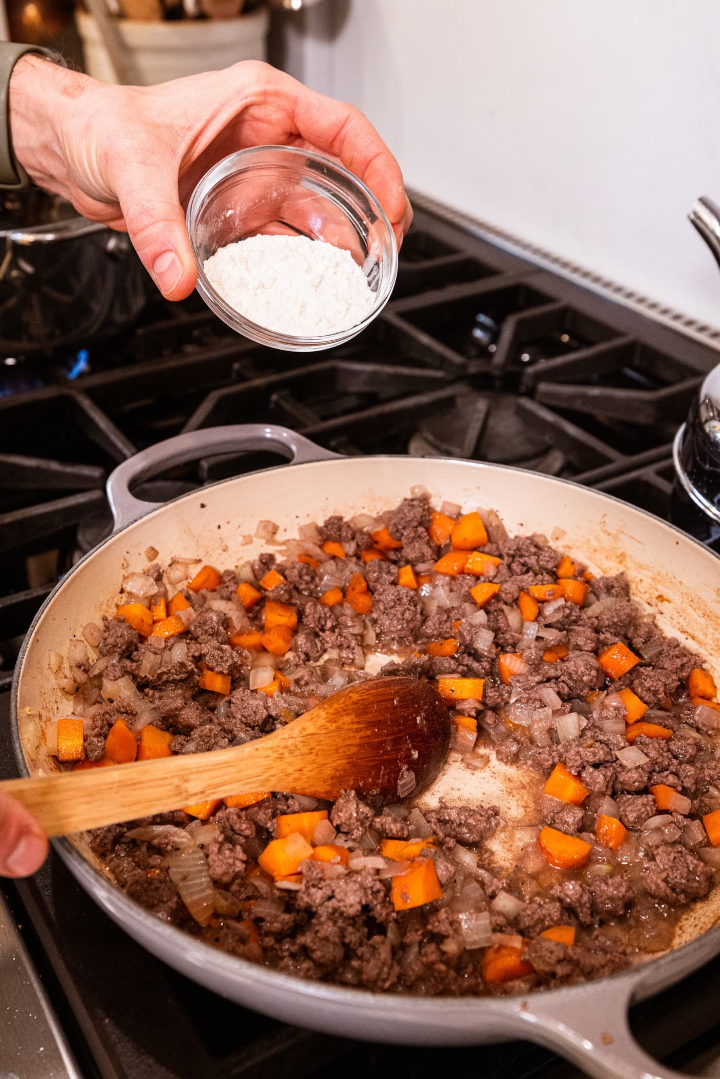 Hand holding small bowl filled with flour about to pour on top of beef and vegetables in large pot.