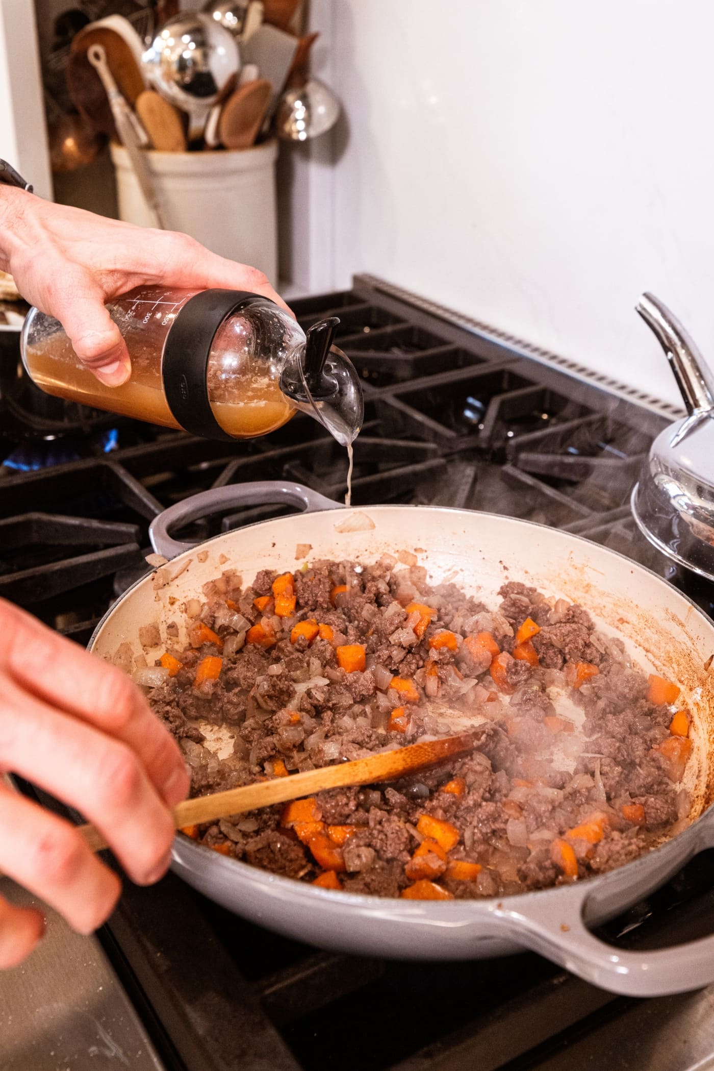 Hand pouring beef stock into a large pot filled with cooking ground beef and vegetable mixture.