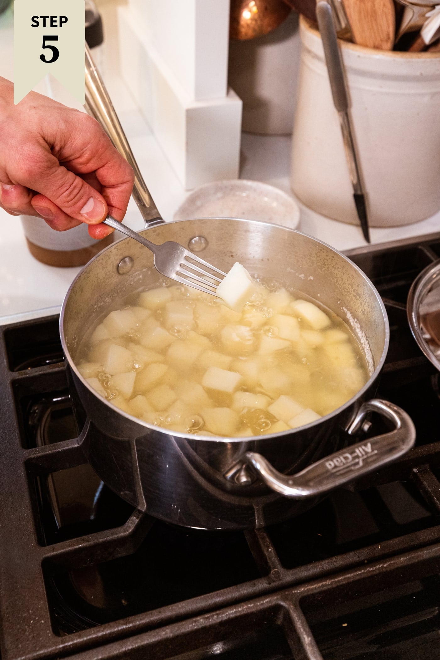 Hand holding fork with piece of diced cooked potato above surface of pot filled with water and cooking potatoes.
