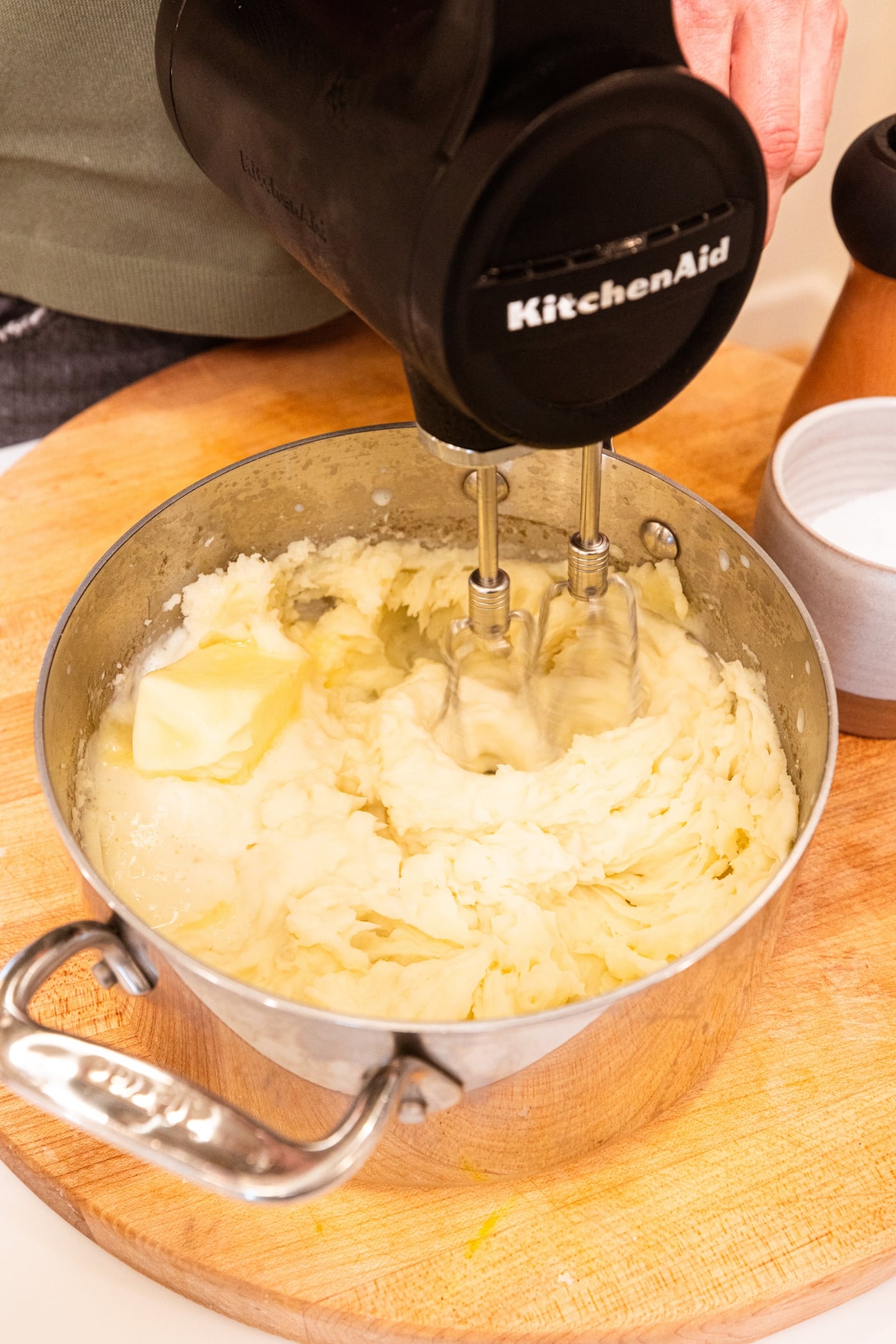 Hand holding black mixer mixing together mashed potato mixture all on wooden surface.