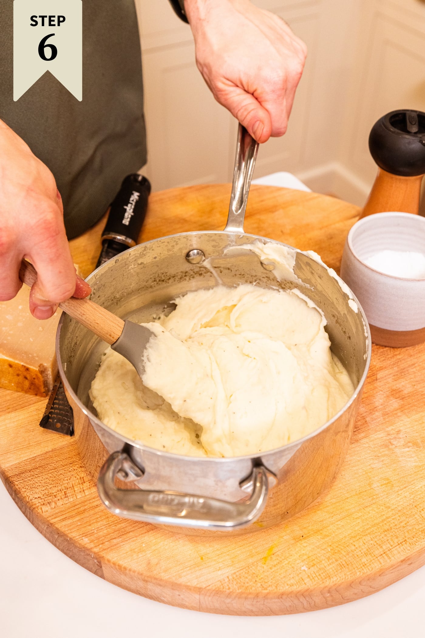 Hand holding spatula mixing together mashed potato mixture in metal pot on wooden surface.