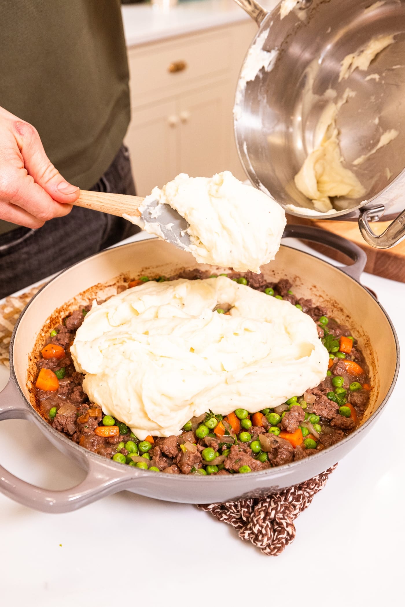 Hand spooning mashed potatoes on top of beef and pea mixture in a large pot after cooking on the stovetop.