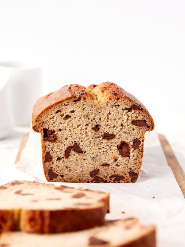 Loaf of chocolate chip banana bread resting on a wooden cutting board with slices resting in front all with white background.