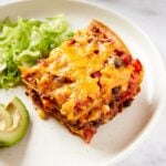 White plate with slice of loaded taco squares casserole showing the multiple layers of tacos and beans mixture with lettuce and avocado.