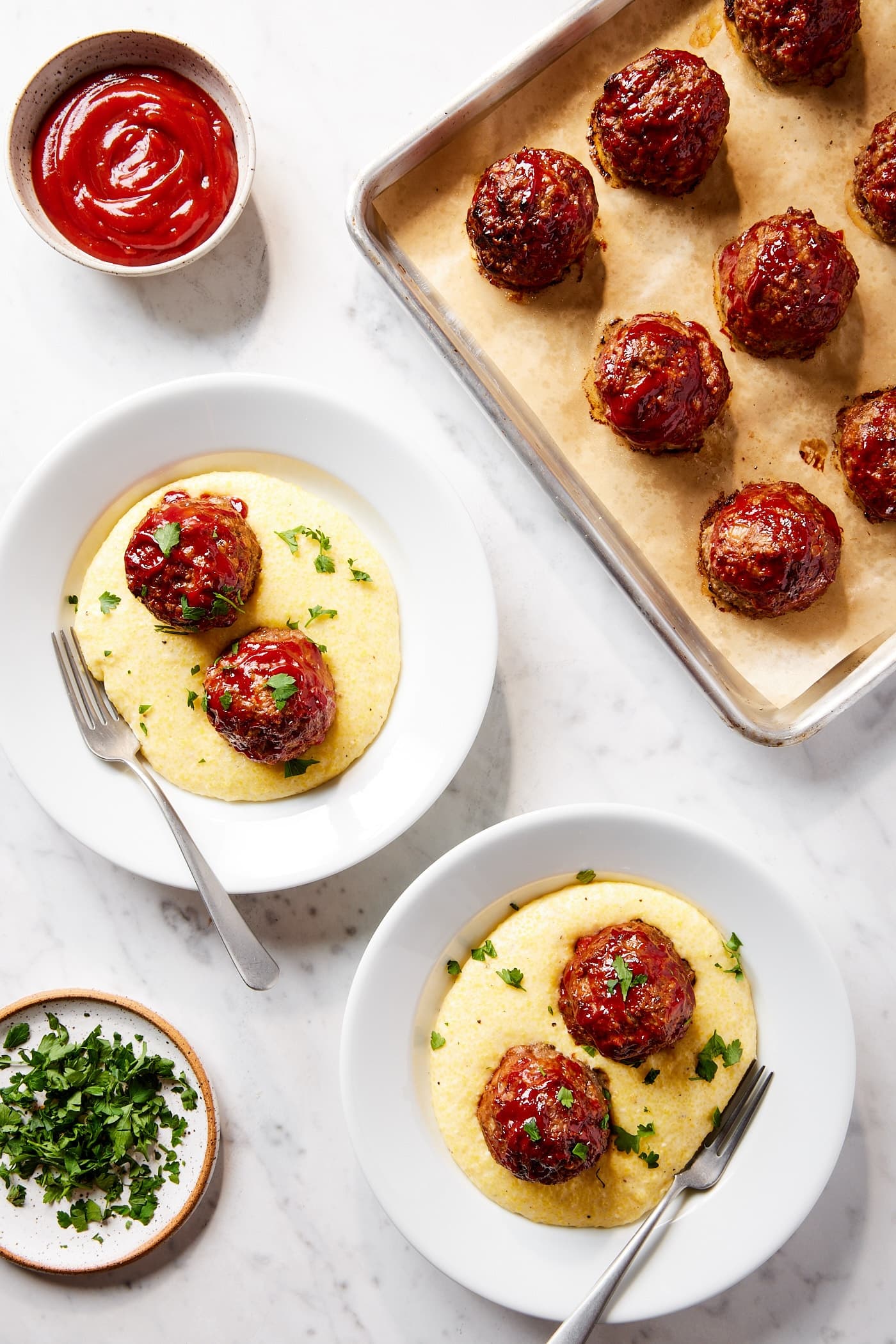 Two white plates on marble surface filled with polenta and meatloaf meatballs with more meatballs on sheet pan.