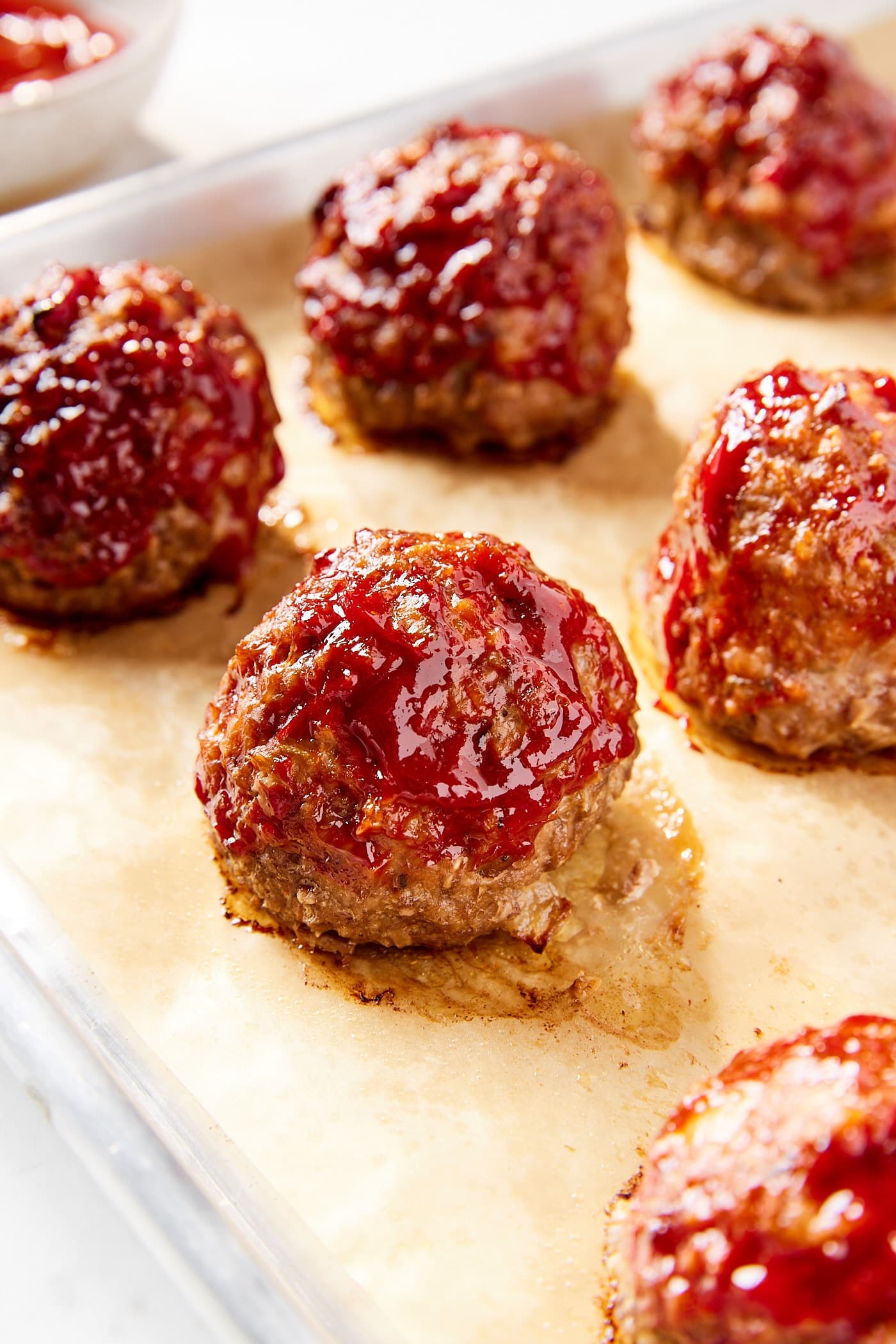 Cooked meatloaf meatballs sitting on parchment-lined baking sheet with ketchup sauce all over the top and sides.