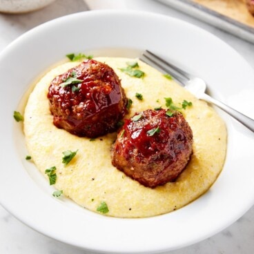 White plate filled with Parmesan polenta with meatloaf meatballs with ketchup sauce on top.