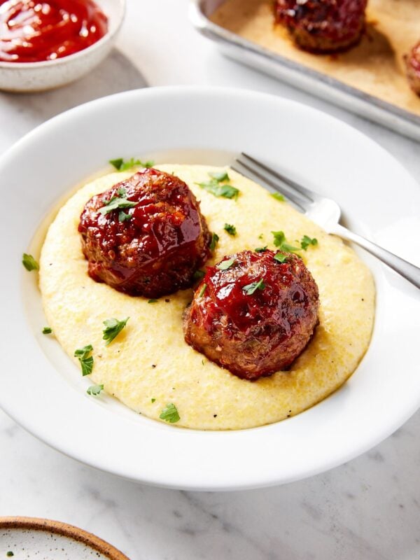 White plate filled with Parmesan polenta with meatloaf meatballs with ketchup sauce on top.