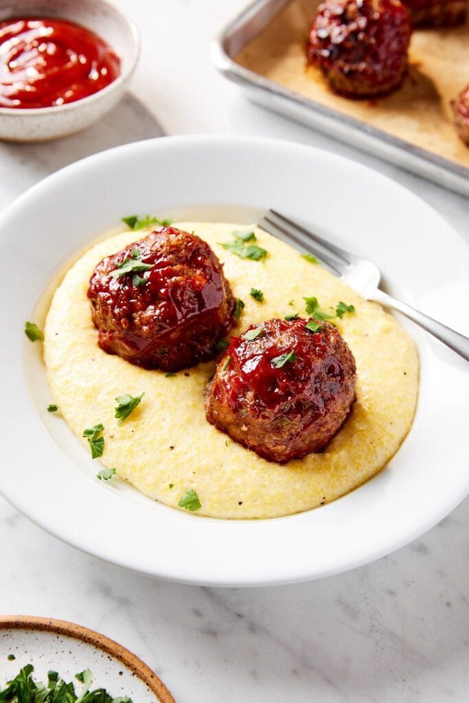 White plate filled with Parmesan polenta with meatloaf meatballs with ketchup sauce on top.