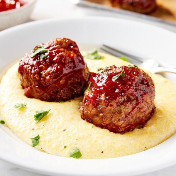 White plate filled with Parmesan polenta topped with two ketchup-covered meatloaf meatballs.