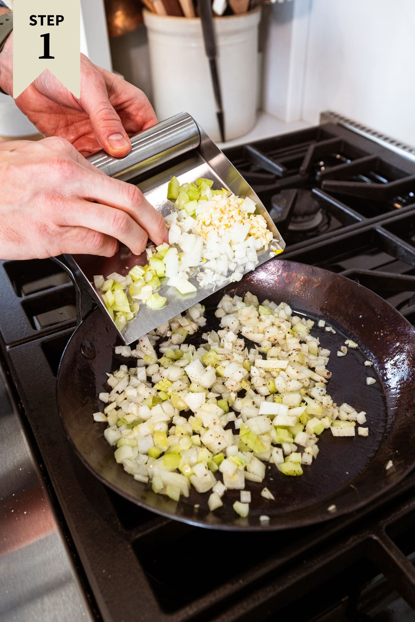 Metal scooper holding diced onion and celery mixture being poured into a black skillet on the stovetop.