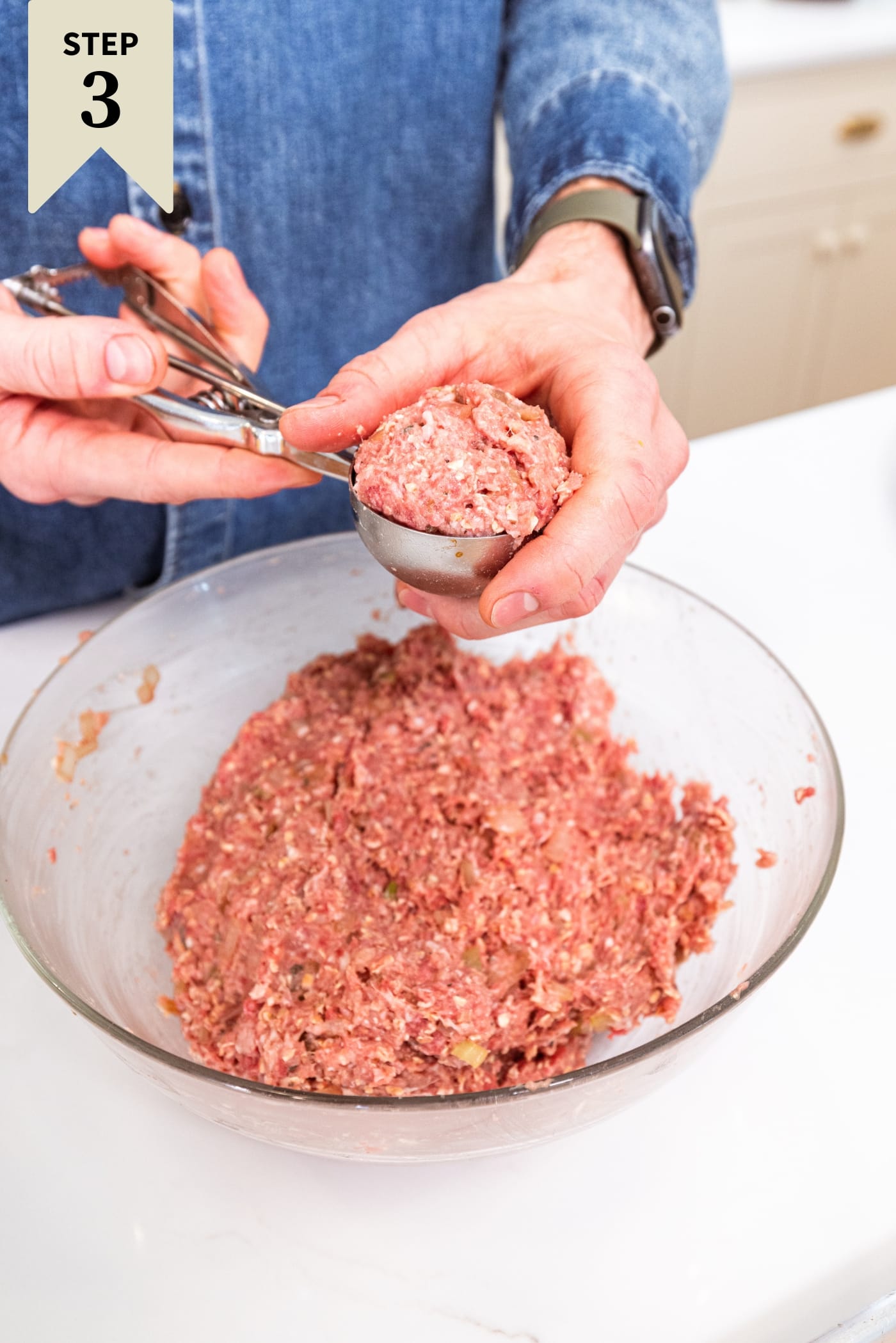 Hand using ice cream scoop to form meatloaf meatballs into meatball shape from glass bowl with the rest of the mixture.