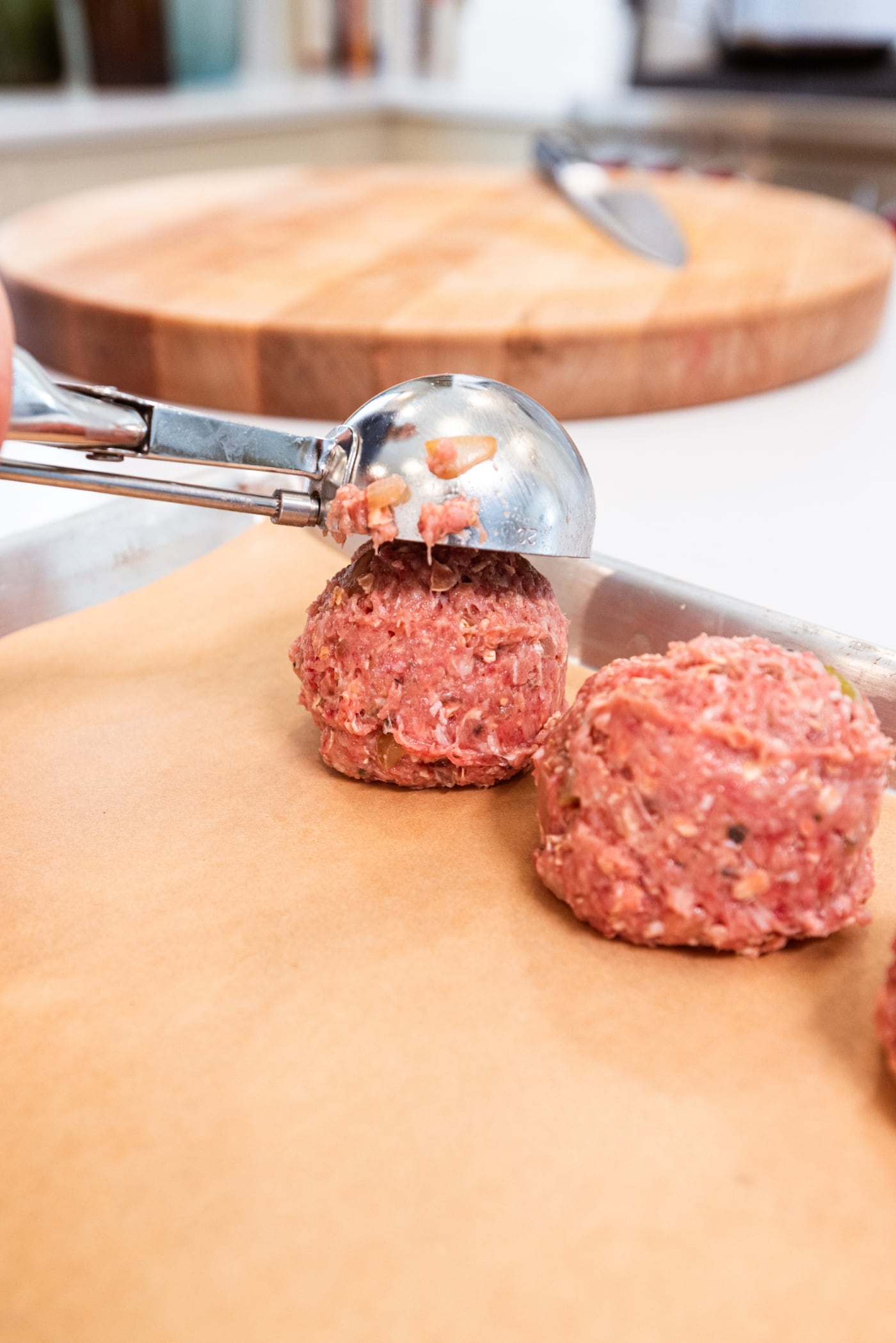 Ice cream scoop dropping a meatloaf meatball on a parchment-lined baking sheet beside another raw meatball.