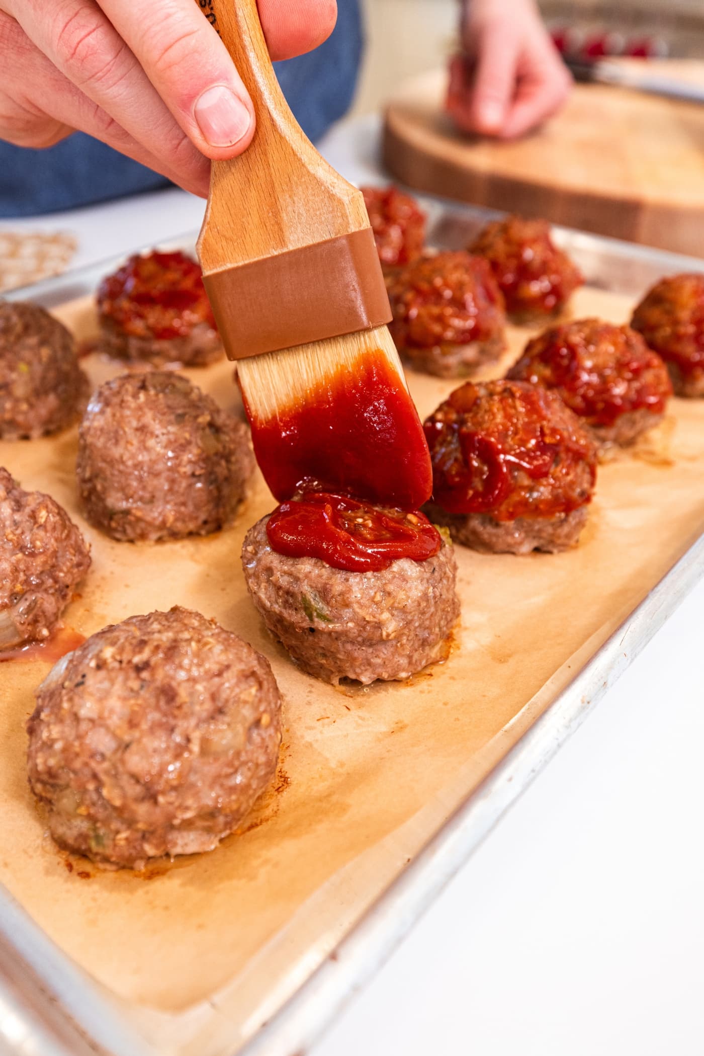 Brush brushing ketchup mixture on top of slightly cooked meatloaf meatballs on a parchment-lined baking sheet.