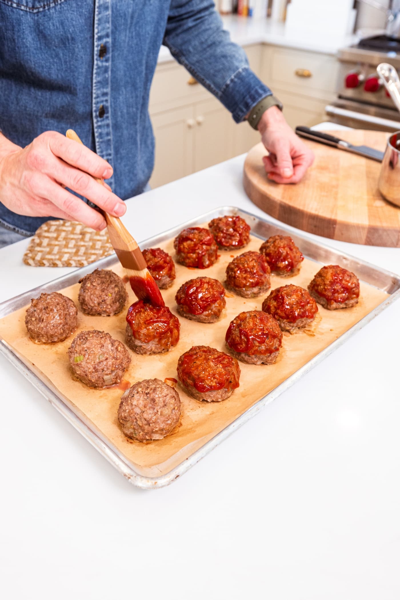 Hand using a brush to brush ketchup mixture on top and sides of meatloaf meatballs on parchment-lined baking sheet.