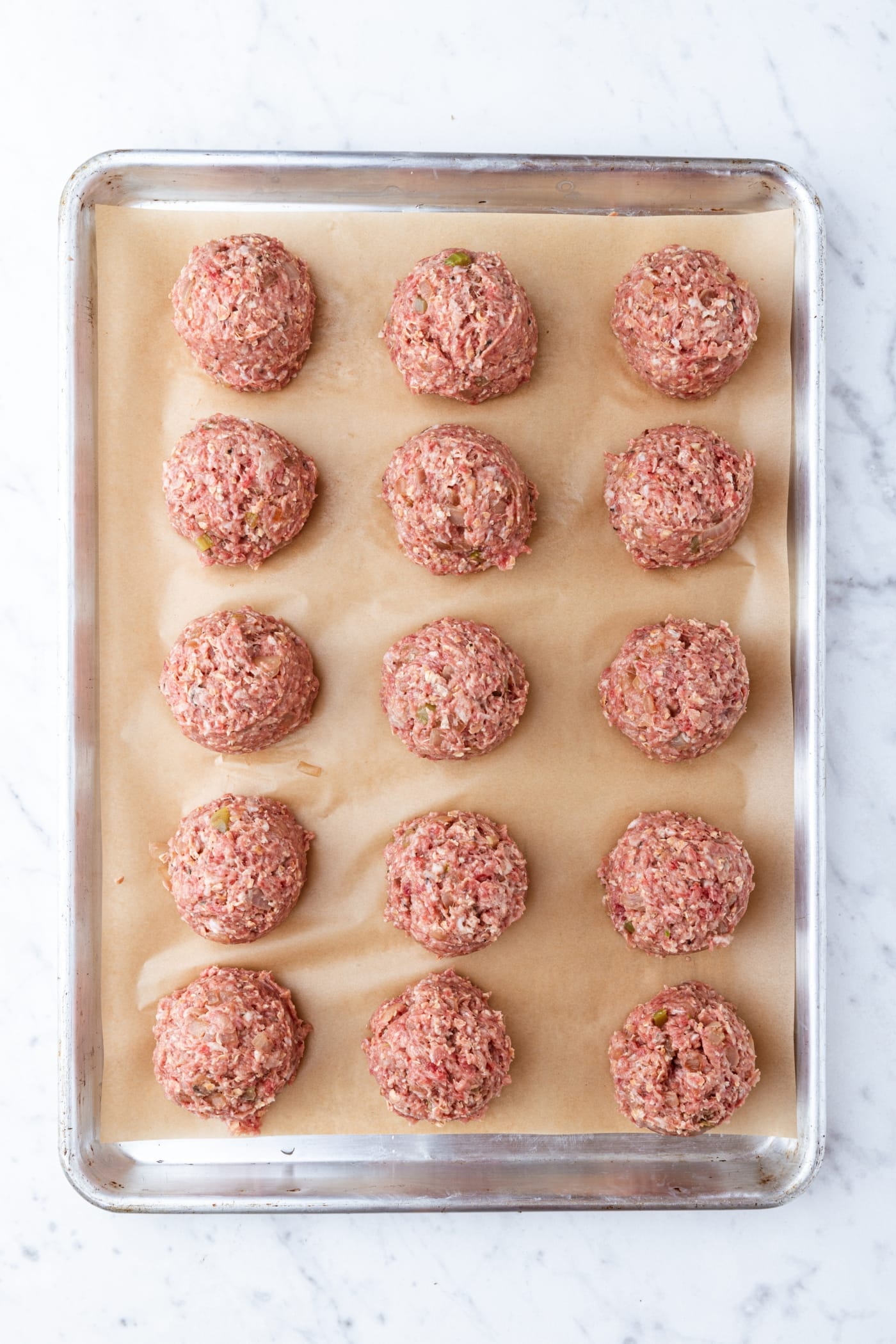 Raw meatballs on a parchment-lined baking sheet on a marble surface before being baked.