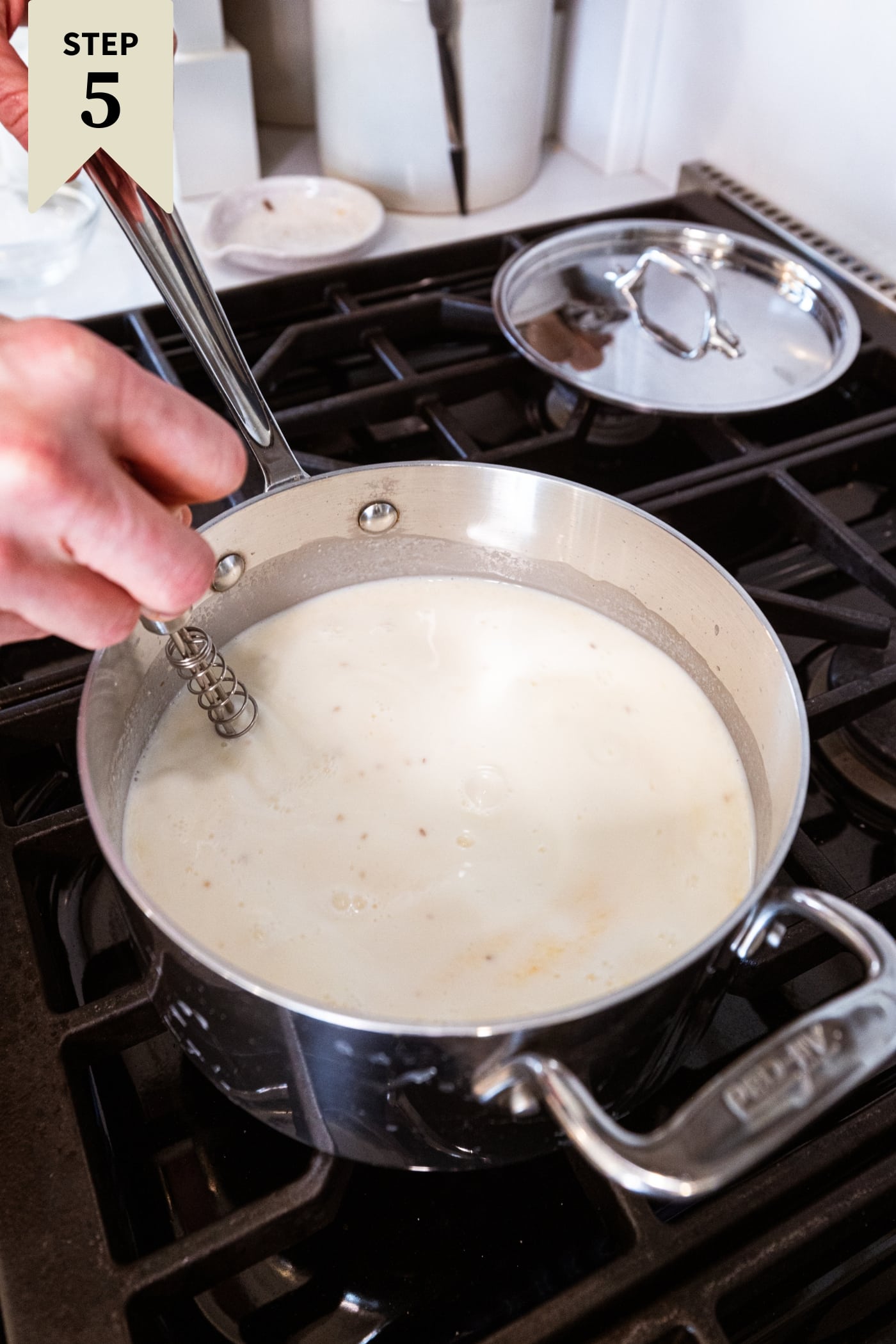 Milky mixture in a metal saucepan set on the heat on a stovetop with hand using a metal whisk to mix everything.