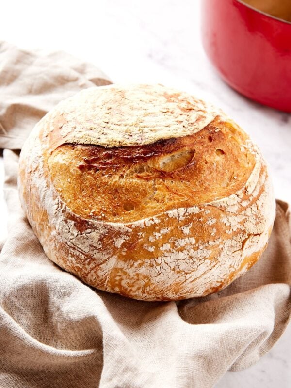 Loaf of baked sourdough bread resting on a piece of cloth with golden crust with red Dutch oven in the background.