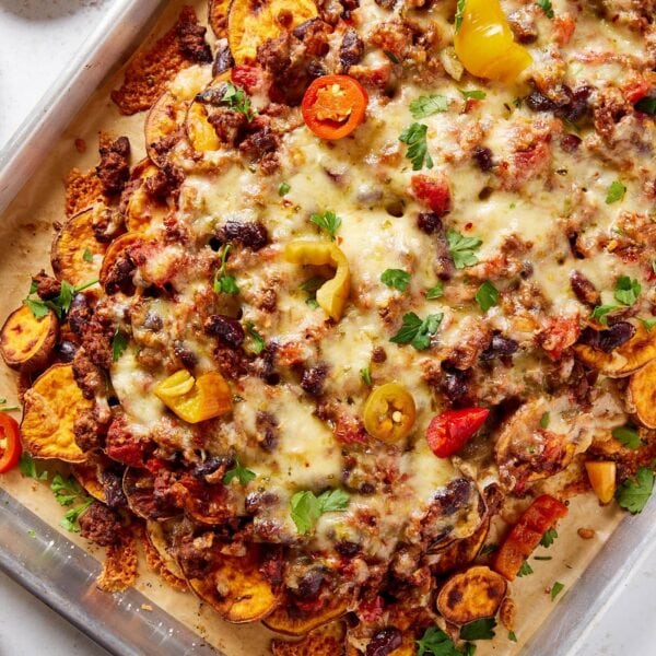 Metal sheet pan filled with layers of sweet potato, chili, and melted cheese along with pickled peppers, more cheese, and slices of avocado.