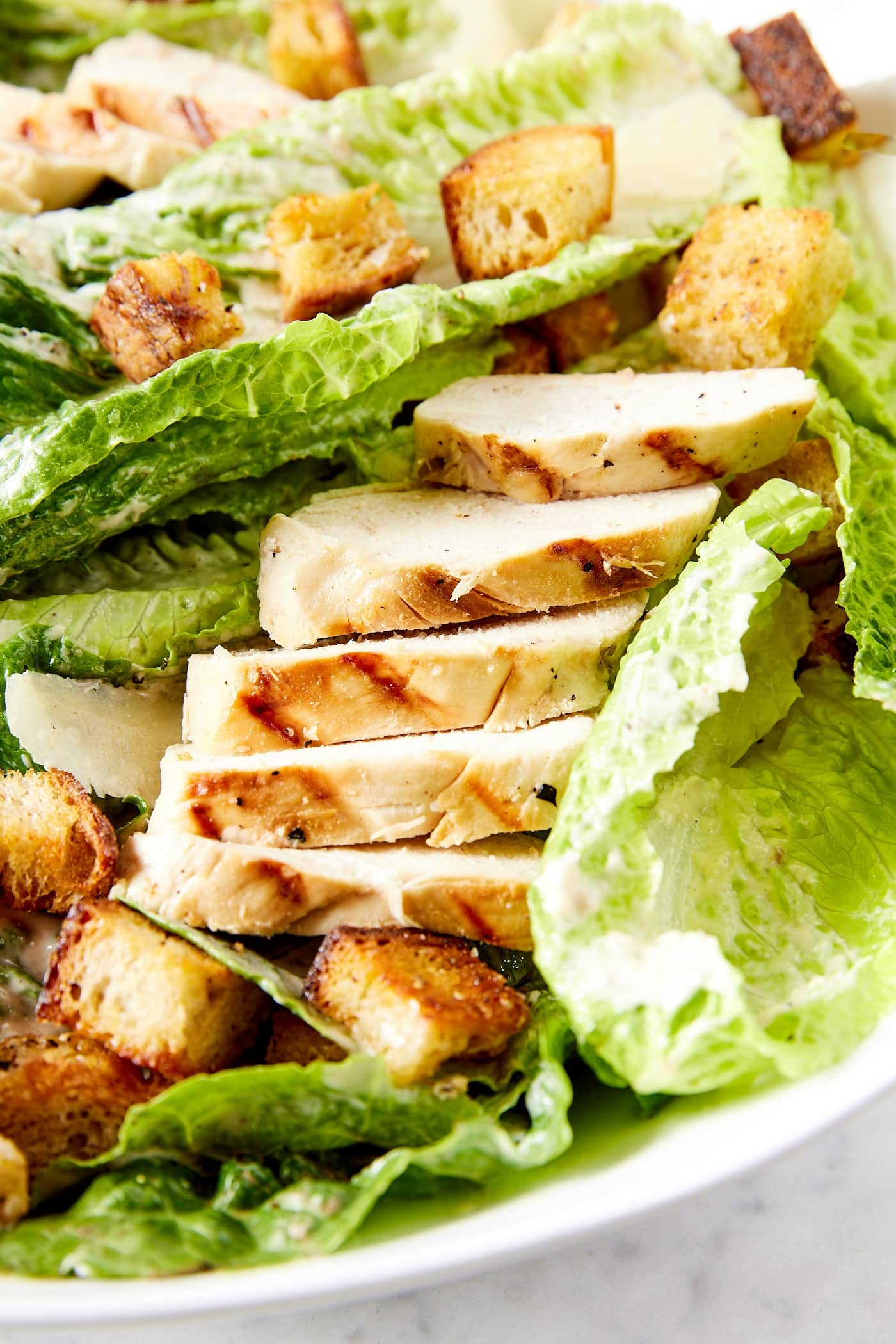 Slices of grilled chicken resting against lettuce and croutons nestled in pieces of lettuce with Caesar dressing on top.