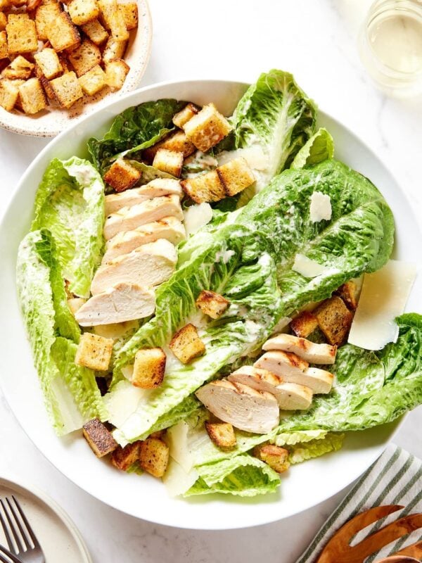 Large white bowl filled with chicken Caesar salad with croutons sprinkled on top with glass of white wine to the side.
