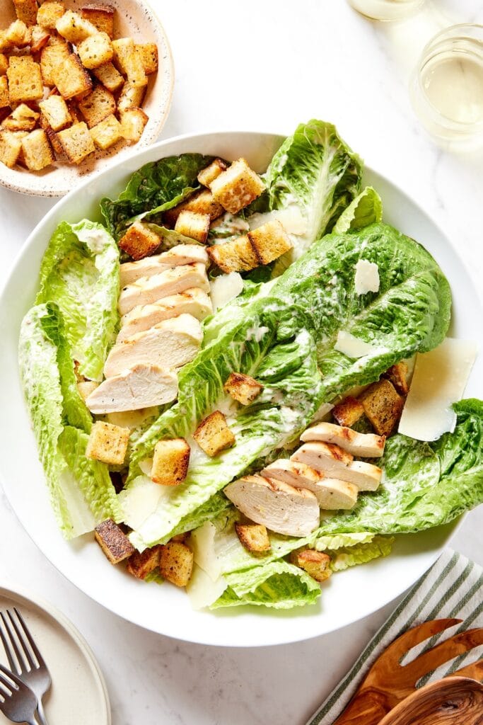 Large white bowl filled with chicken Caesar salad with croutons sprinkled on top with glass of white wine to the side.
