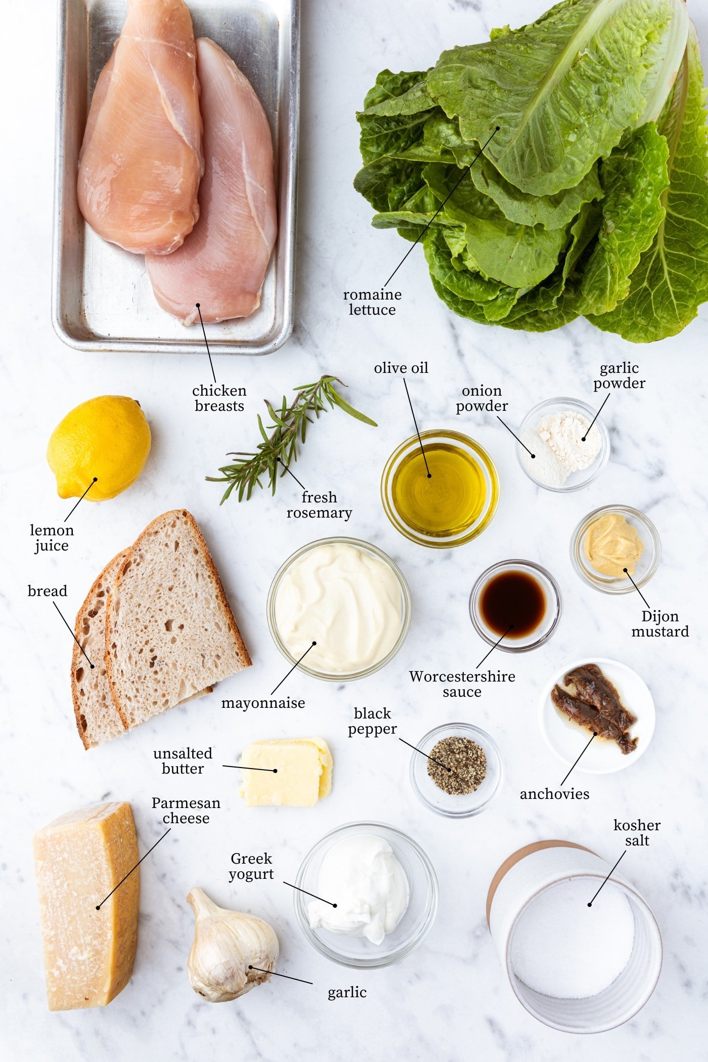 White marble surface filled with all ingredients needed for chicken Caesar salad including lettuce, bread, mayonnaise, spices, and more.