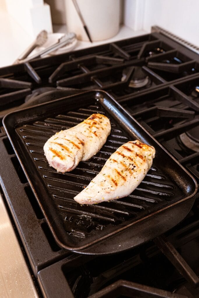 Two chicken breasts cooking on a grill on a stovetop with grill marks on top.