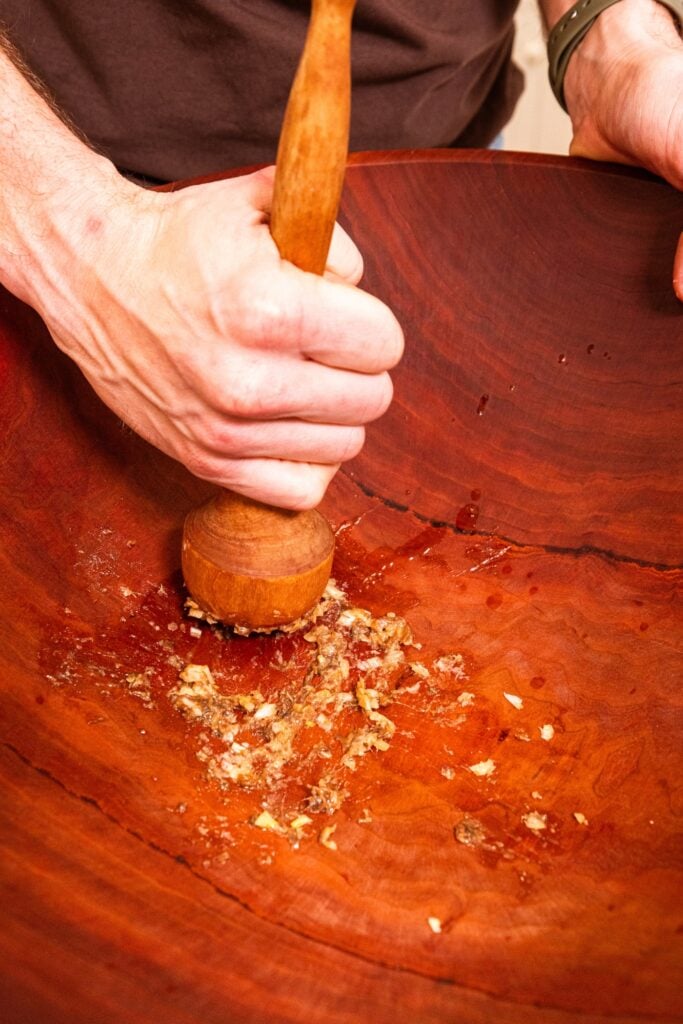 Hand using mallet to crush together garlic and anchovies in large wooden bowl for homemade Caesar dressing.