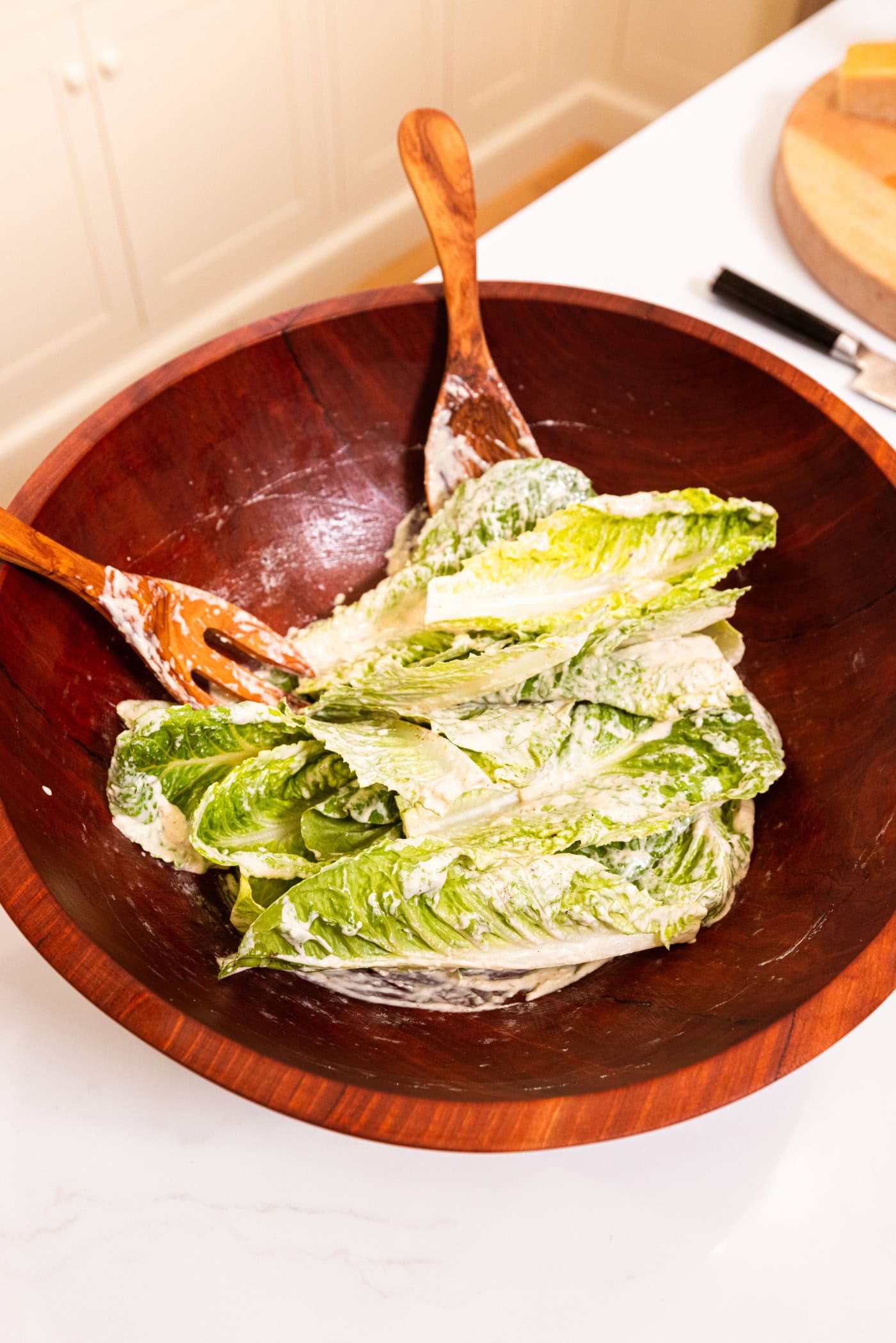 Pieces of lettuce being tossed in large wooden bowl with Caesar dressing with two wooden salad tongs.
