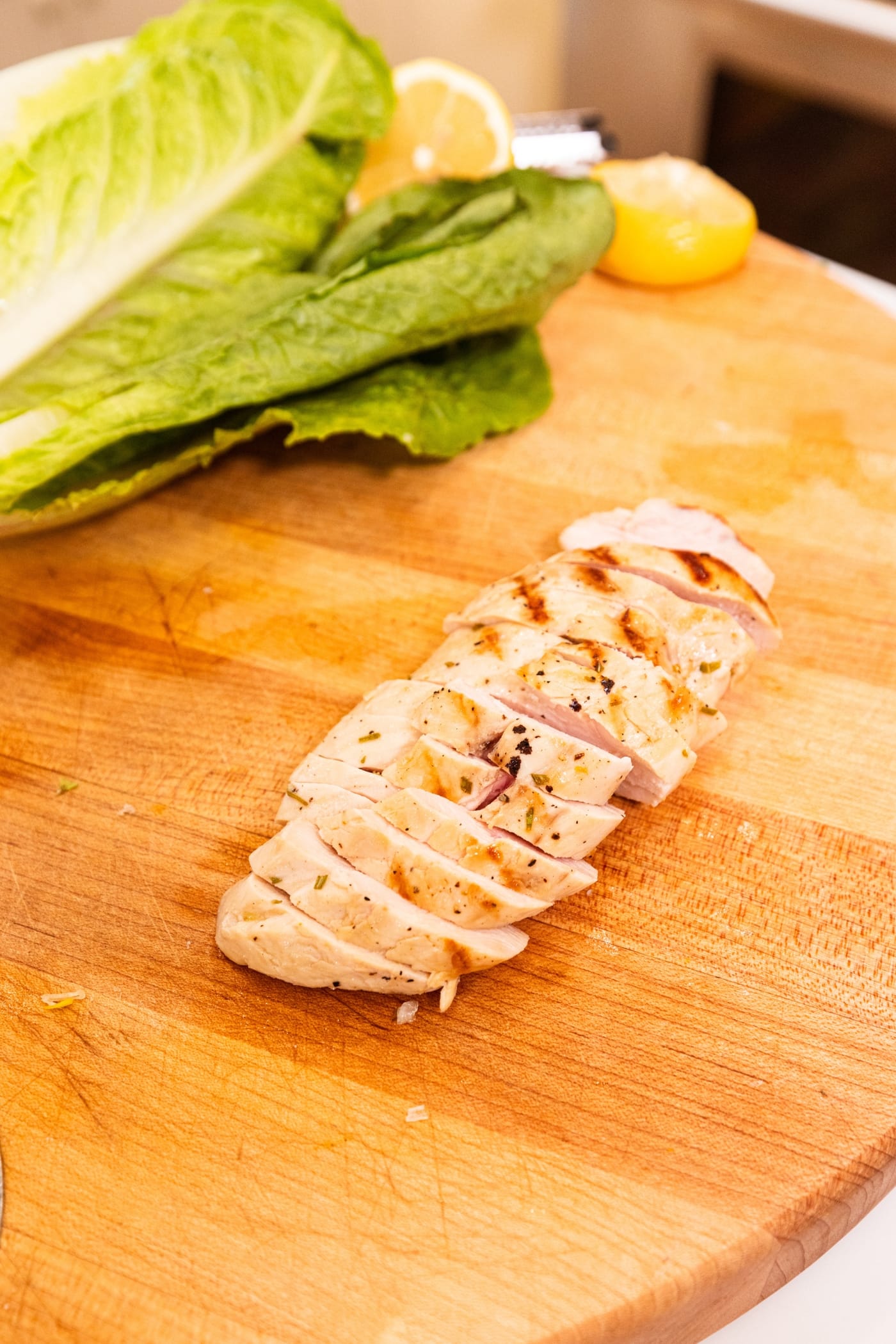 Grilled chicken sliced on into thin pieces in wooden cutting board with lettuce and a lemon in the background.
