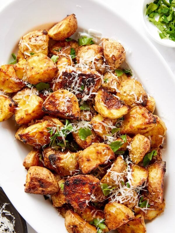 Oval serving dish filled with rosemary roasted potatoes topped with cheese and fresh herbs with crispy exterior texture.