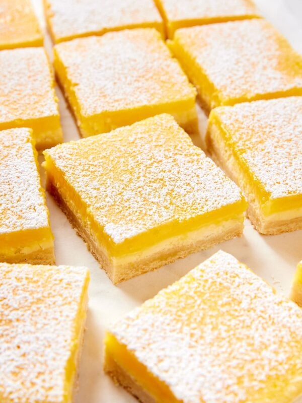 Lemon cheesecake bars resting on a white parchment surface dusted with powdered sugar showing each layer of the bar.