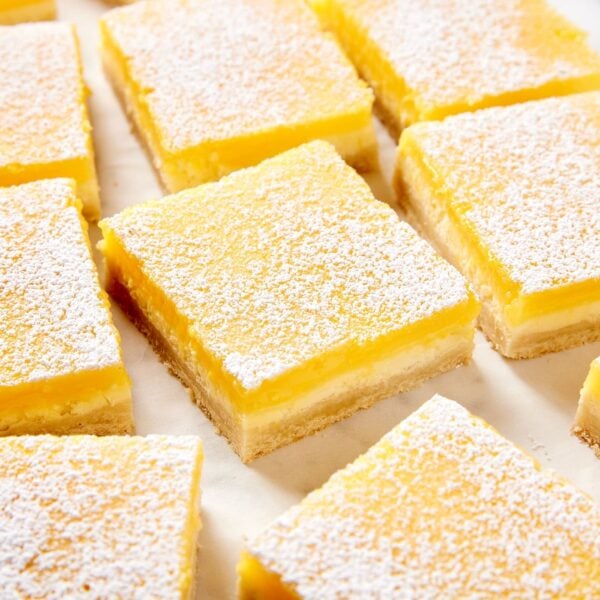 Squares of lemon cheesecake bars sitting on piece of white parchment dusted with powdered sugar.