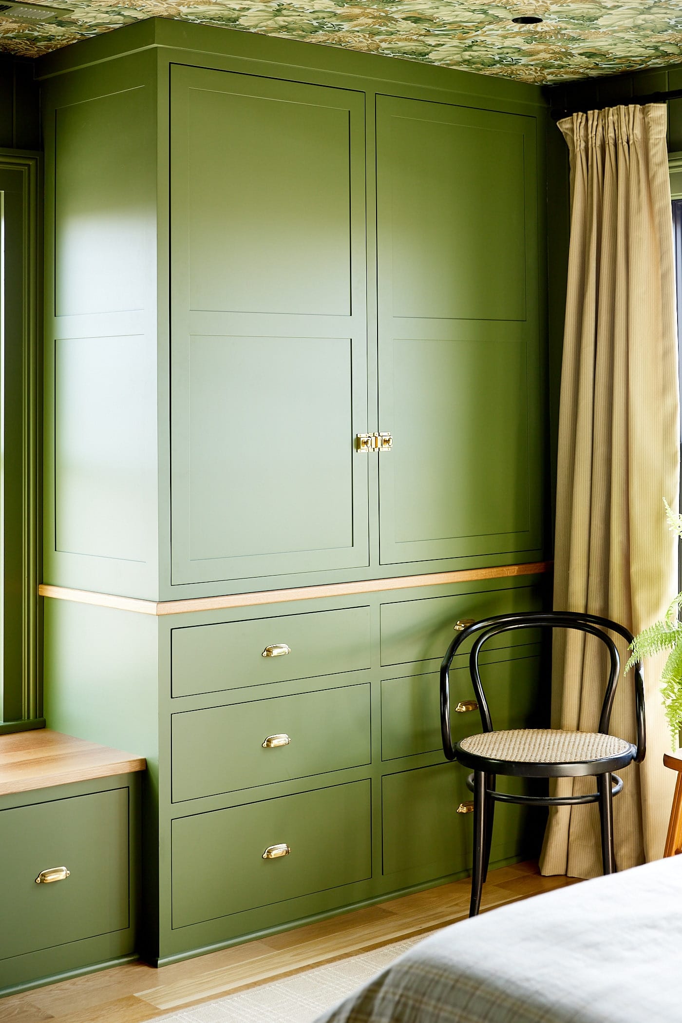 Green colored cabinets with doors and drawers with chair sitting in front along with window seat to the side all with white oak floors.
