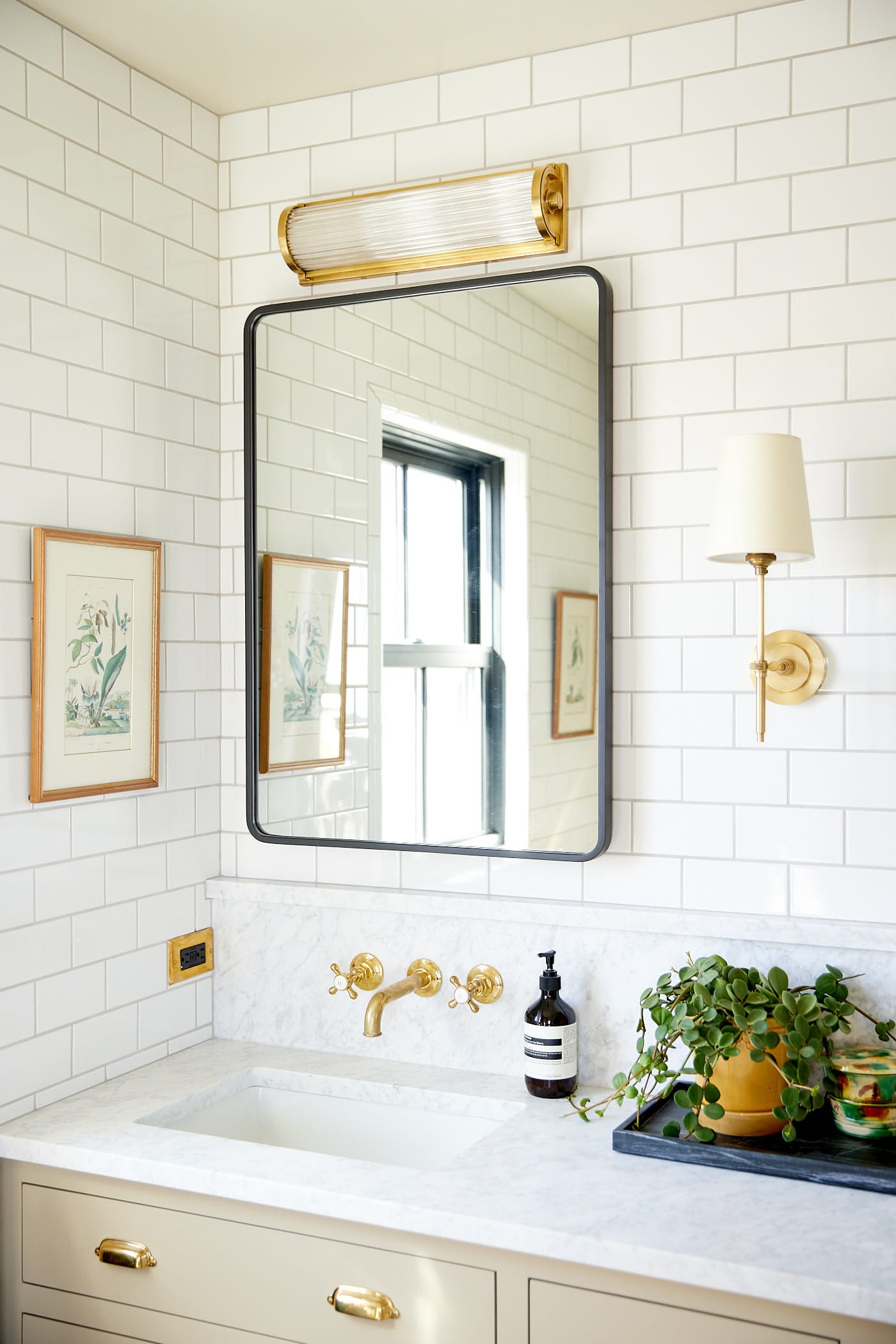 Subway tile covered walls in a bathroom with mirror on wall with light on top with small painting to the side and brass fixtures for sink basin.