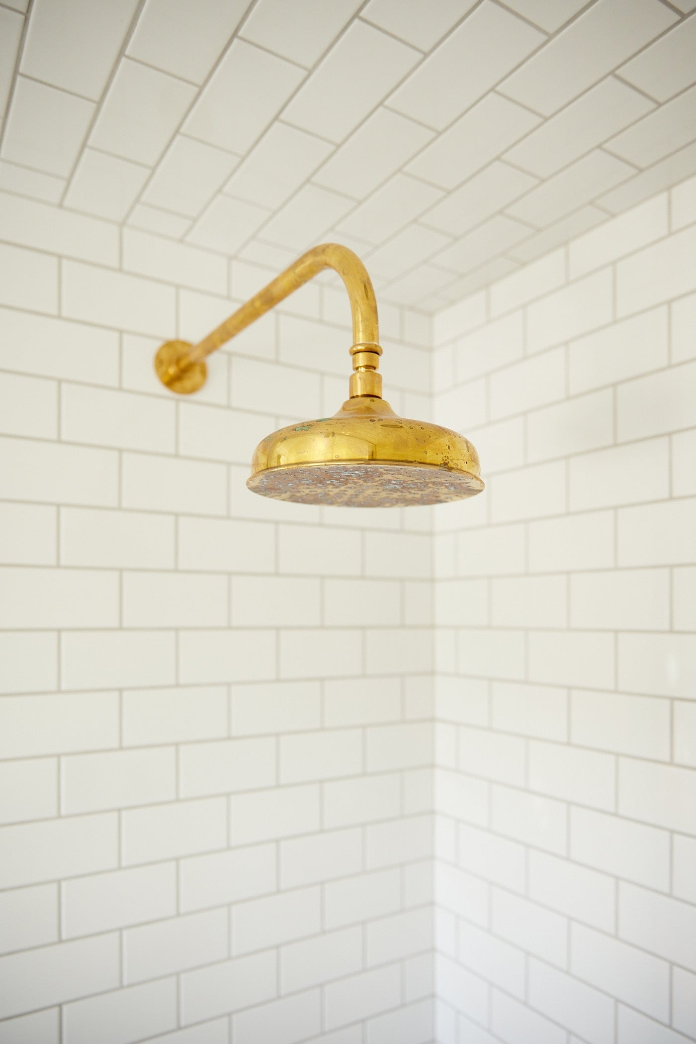 Brass shower head with age spots all over in shower filled with subway tile on all walls.