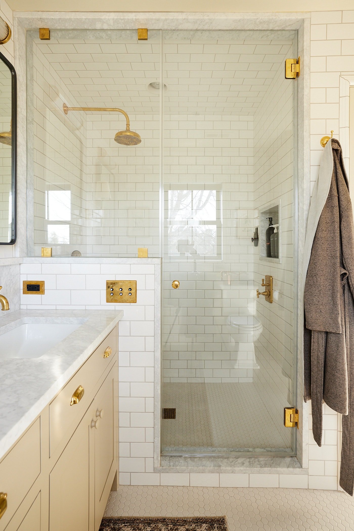 Glass and tile shower with brass fixtures with door right beside a khaki colored cabinet with mirror and a robe hanging to the side.