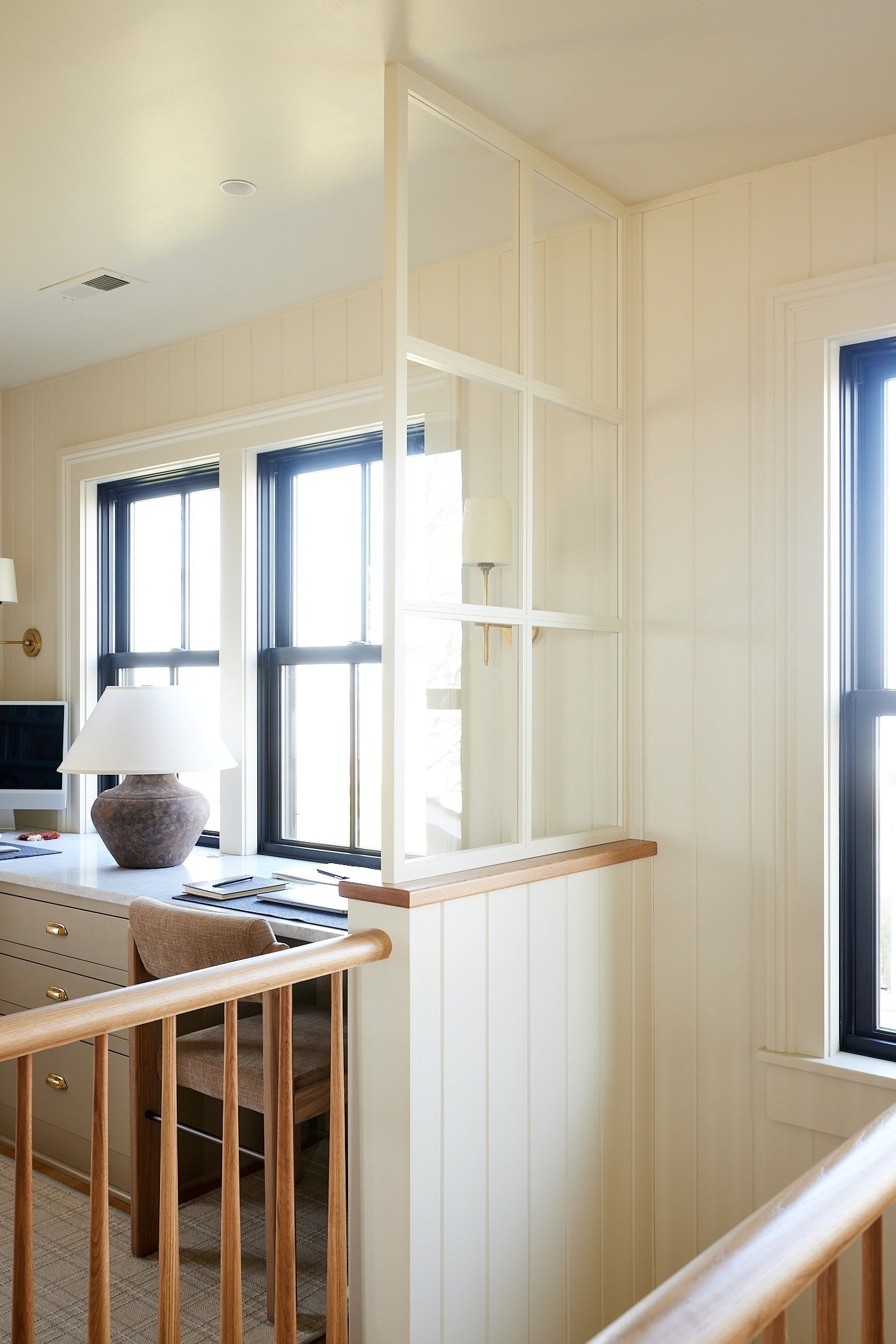 Half wall with interior glass window on top with railing extending along the space for stairwell with desk on the other side.