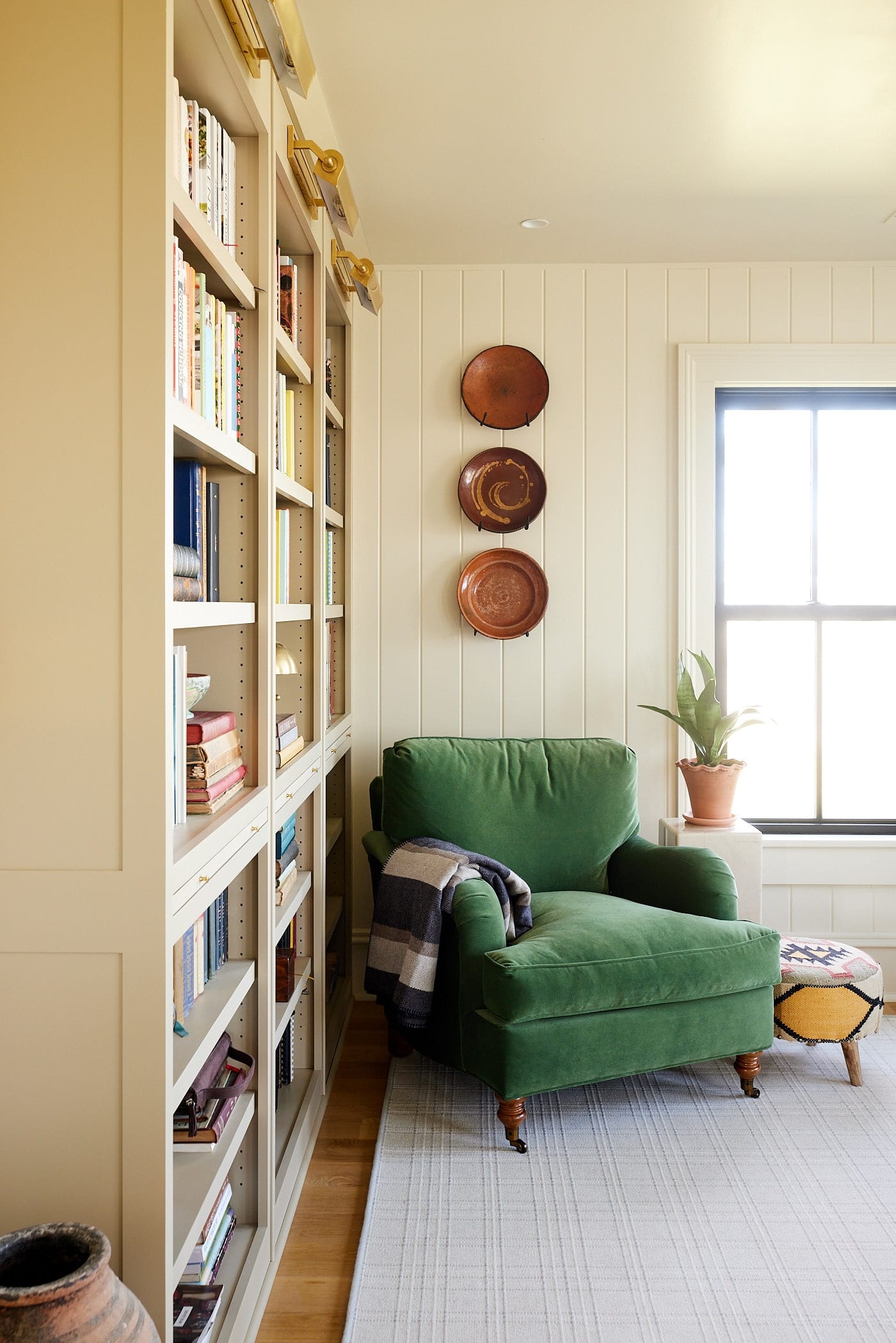 Green comfy chair sitting in front of bookcases filled with cookbooks with window to the side and red plates nailed to the wall.