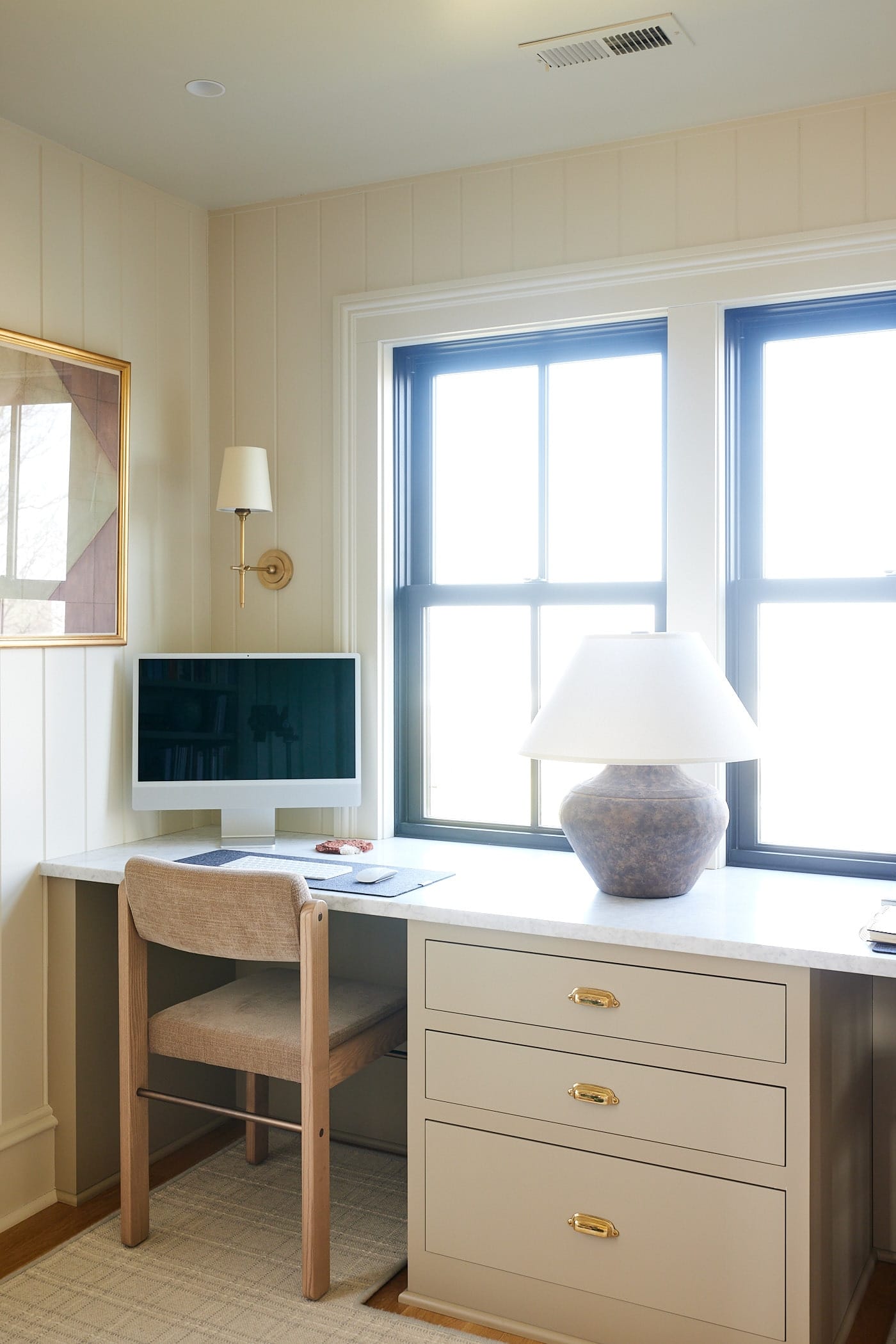 Large built-in desk with marble countertop with computer sitting on top and desk chair in open cubby with lamp all in front of two large windows.