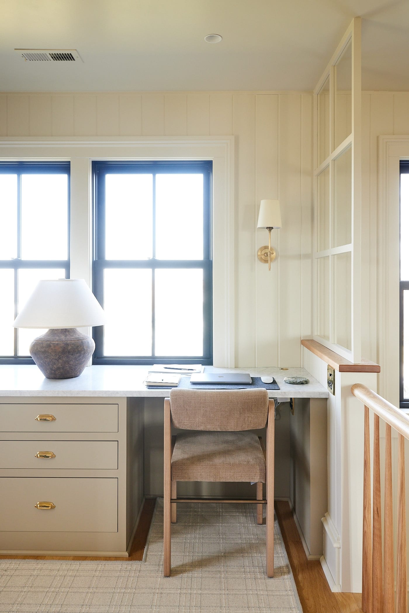 Desk space with chair in front of two large windows with desk lamp and drawers to the side along with white oak railing.