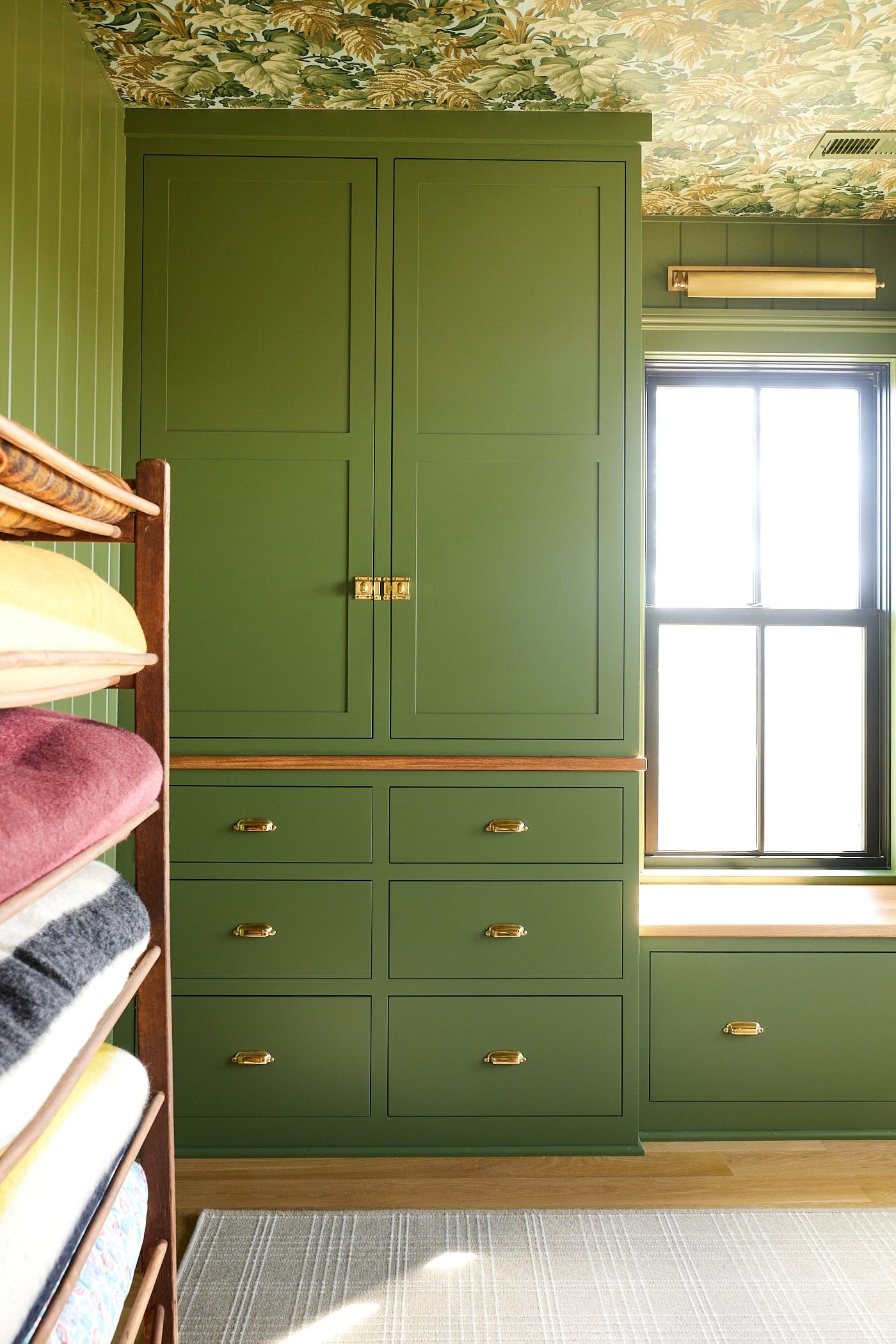 Green colored cabinet acting as a closet for a bedroom with brass hardware with window seat to the side and blanket rack to the front.